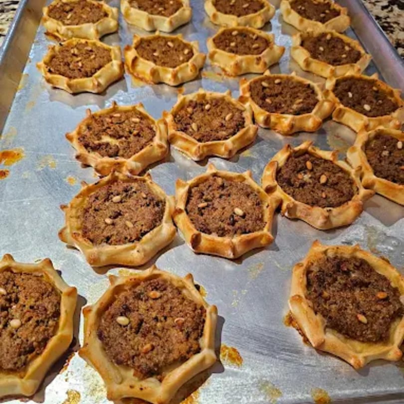 A baking sheet filled with numerous savory meat pies, each topped with pine nuts.