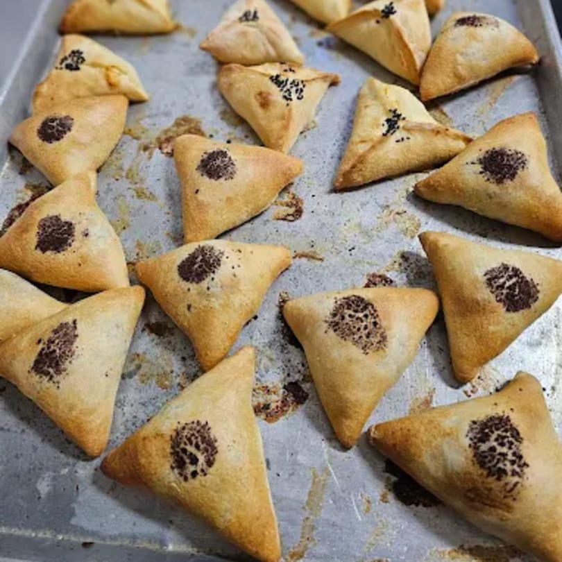 A baking sheet filled with golden-brown, triangular pastries, some topped with dark seeds.