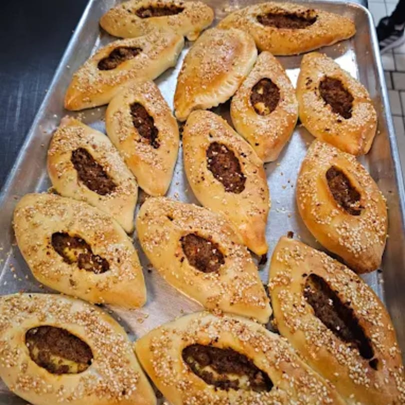 A metal tray filled with freshly baked, boat-shaped pastries topped with sesame seeds and a savory filling.